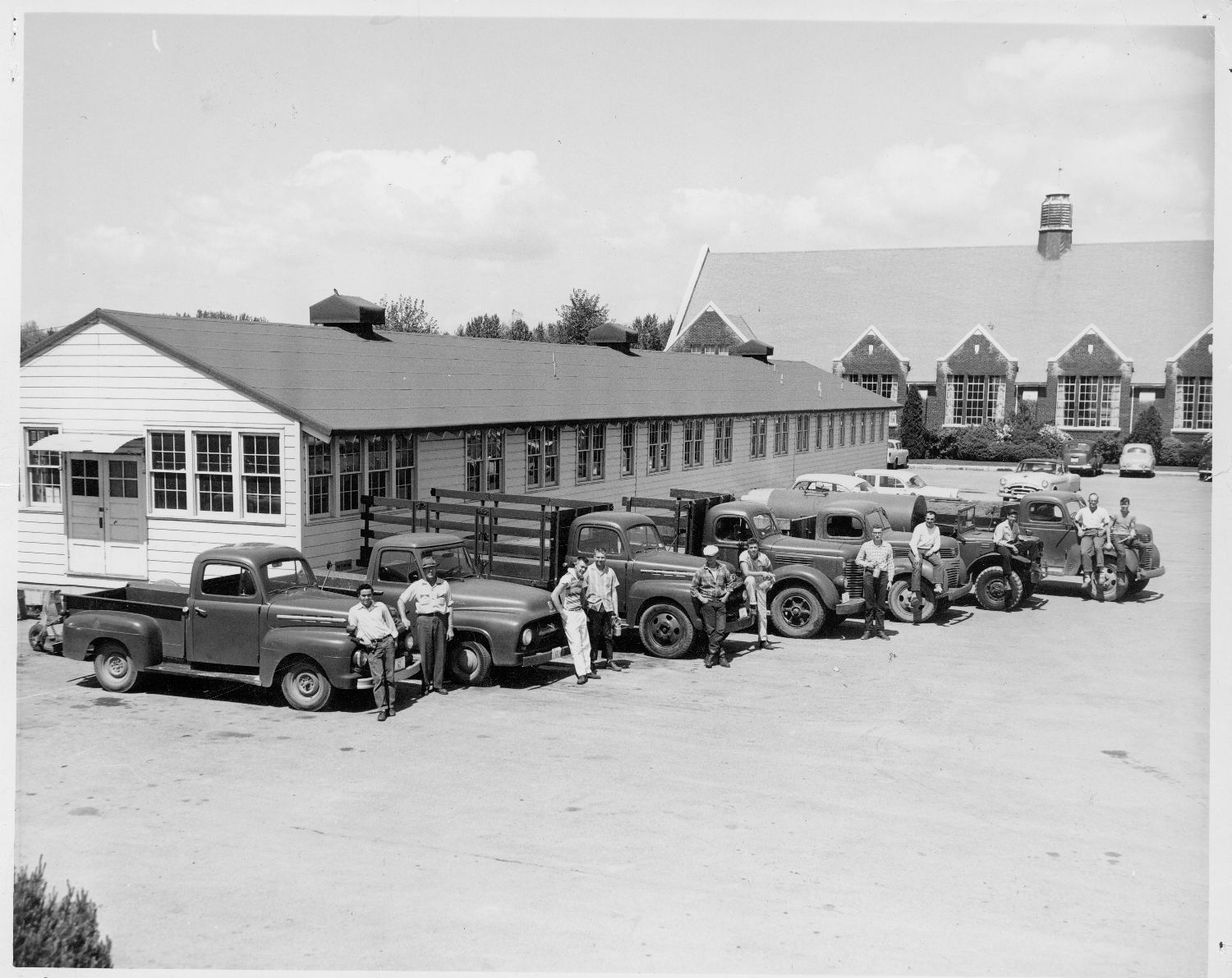 Men pose with a line of trucks. The men are holding paint guns and hammers; they are most likely students of the vocational program. Behind the line of trucks is a repurposed World War II barracks building.