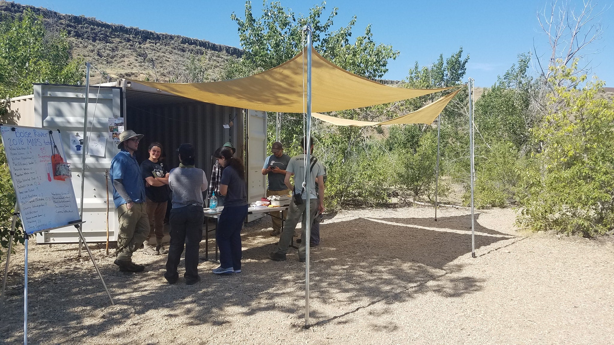 a group of people stand underneath a shade cloth stretched between metal poles