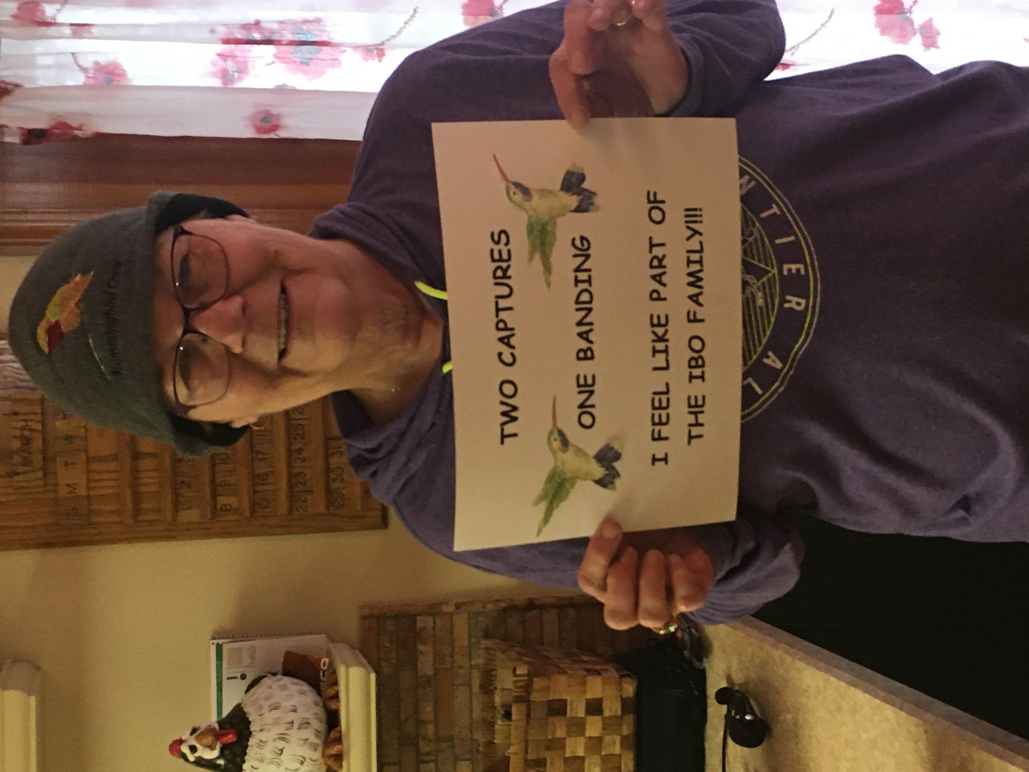 a woman with thinning hair wears a warm knit hat with a hummingbird embroidered on it. she's holding a sign with hummingbird clipart on it that reads "two captures one banding. I feel like part of the IBO family!!!"