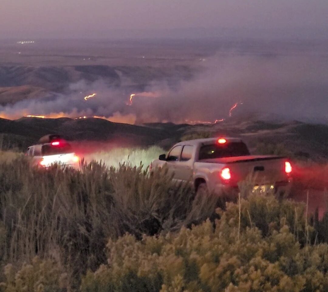 pickup trucks with bright red tail lights glowing in the early morning light head down the road among sagebrush and rabbitbrush. The foothills on the near horizon are completely engulfed in flame with a glowing orange line of fire that is advancing uphill