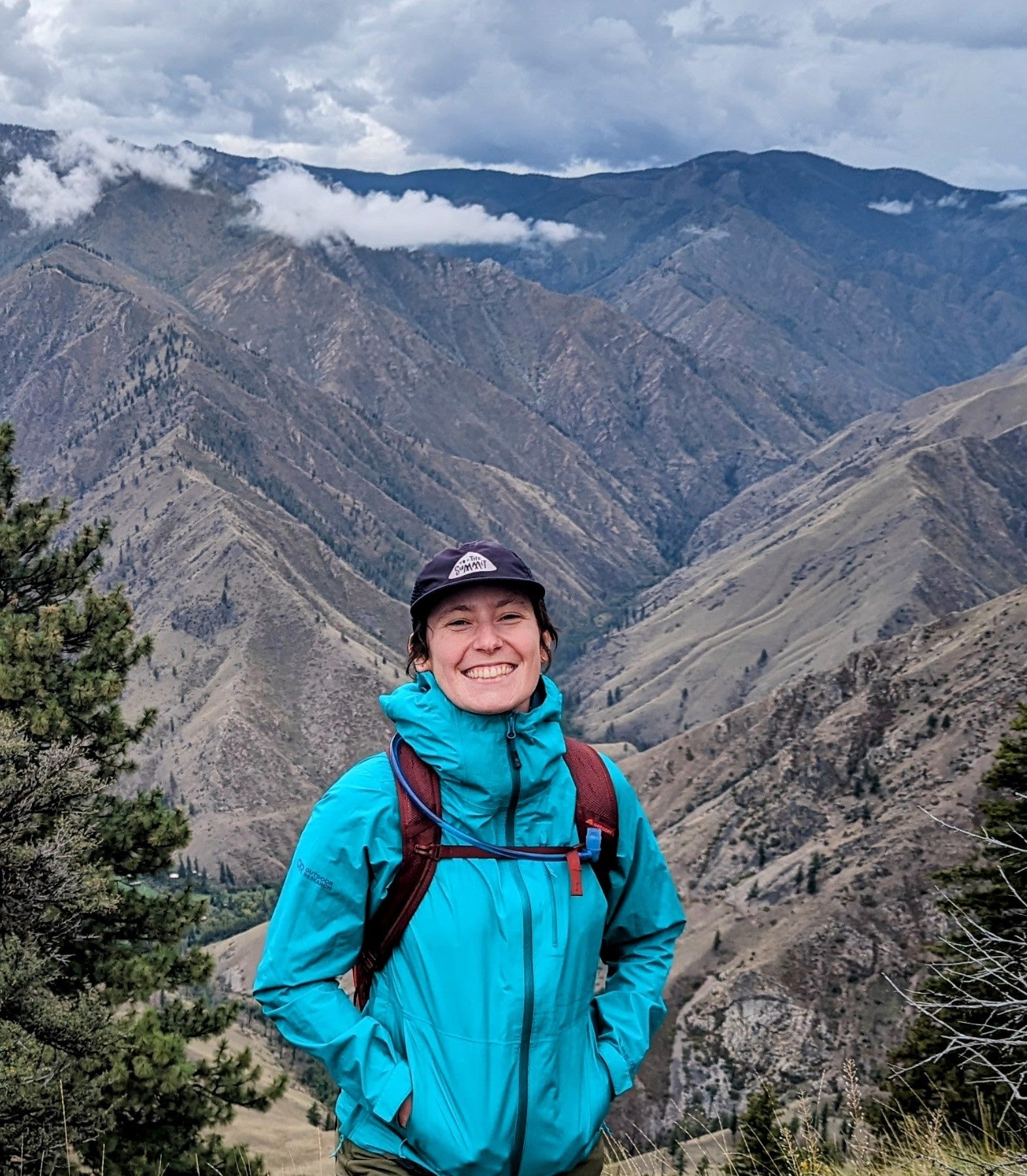 Young woman poses in front of mountains and trees.