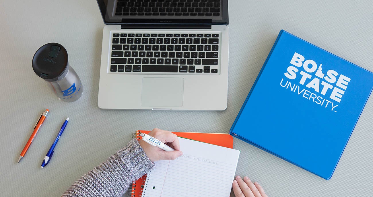 student writing in a notebook, a laptop and office supplies on the table in front of them
